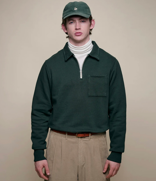 Person wearing a green pullover, beige pants, and a green cap on a beige background