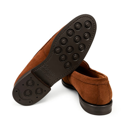 A pair of brown suede slip-on shoes shown on a white background, with one shoe turned to display the black rubber outsole featuring circular tread patterns and the brand name 'Gustavelli' embossed on the heel.