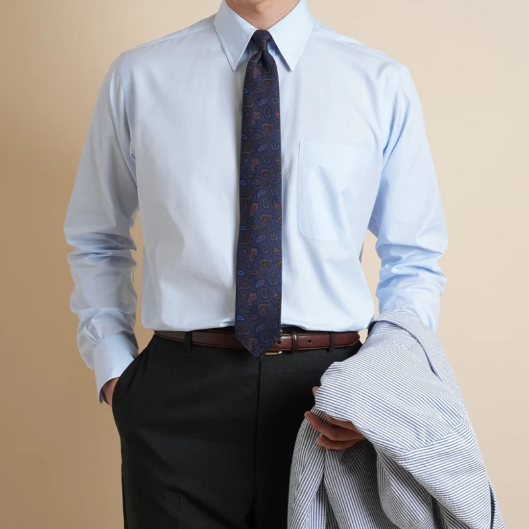 Man wearing a light blue dress shirt, dark tie, black pants, and holding a gray jacket against a beige background