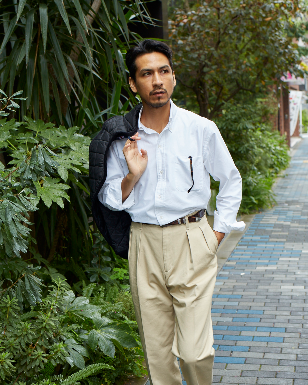 Man in a white shirt and beige pants standing outdoors with greenery around
